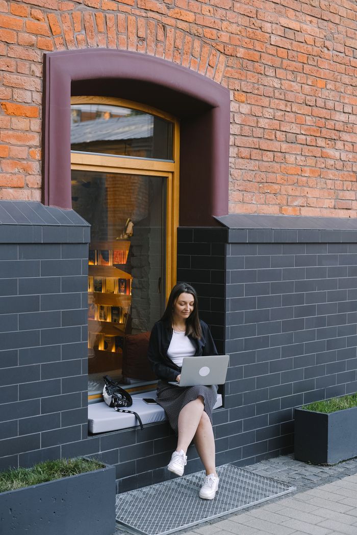 about-04 Woman sitting outside a brick building, working on a laptop, embracing a digital nomad lifestyle.