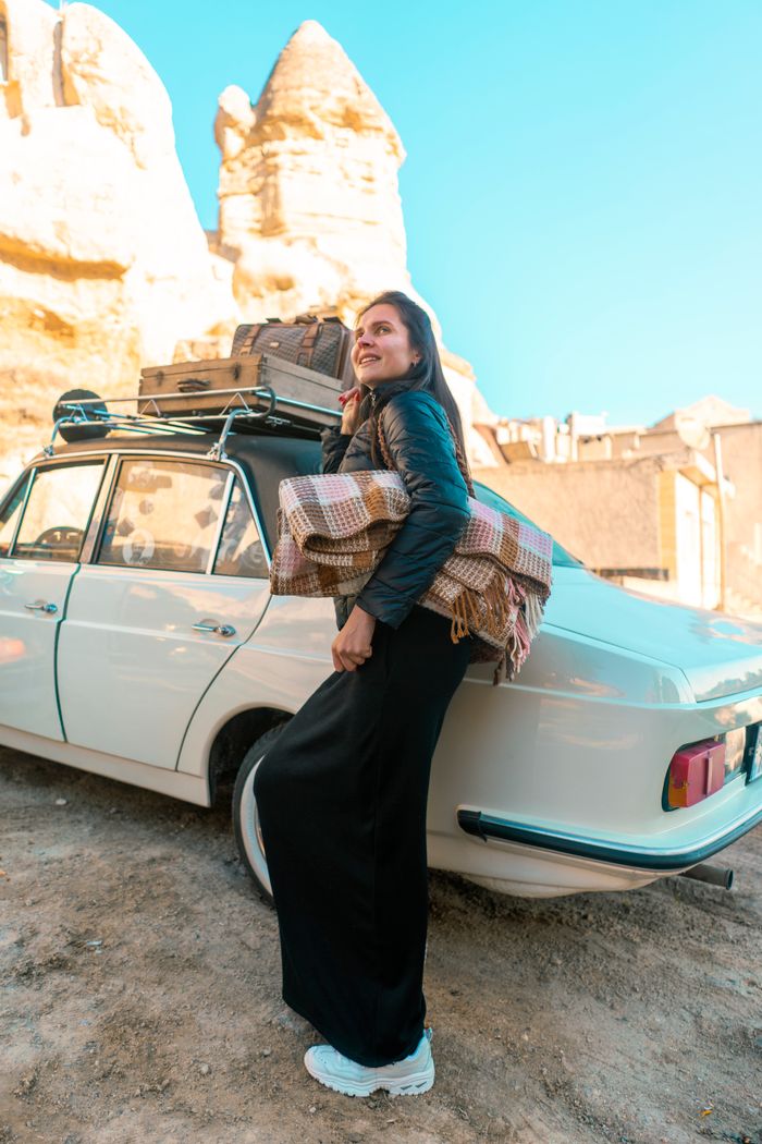Smiling woman leaning on a vintage car with rocky formations in Cappadocia, Turkey.