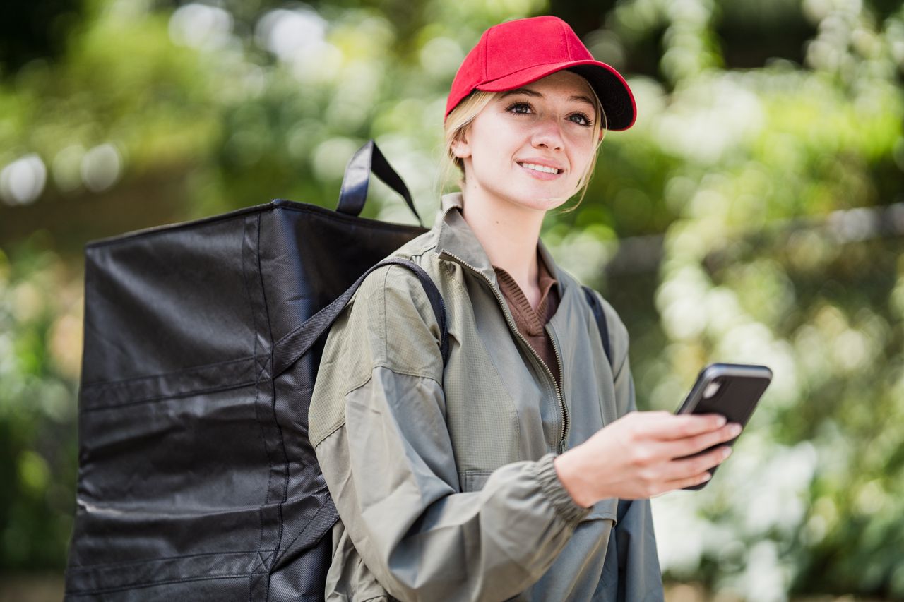 menu-03 Female courier with smartphone using an app outdoors, wearing a red cap and green jacket.