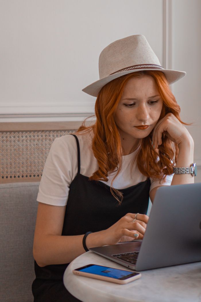 Redhead woman wearing a hat, focused on laptop work with a smartphone on the table.