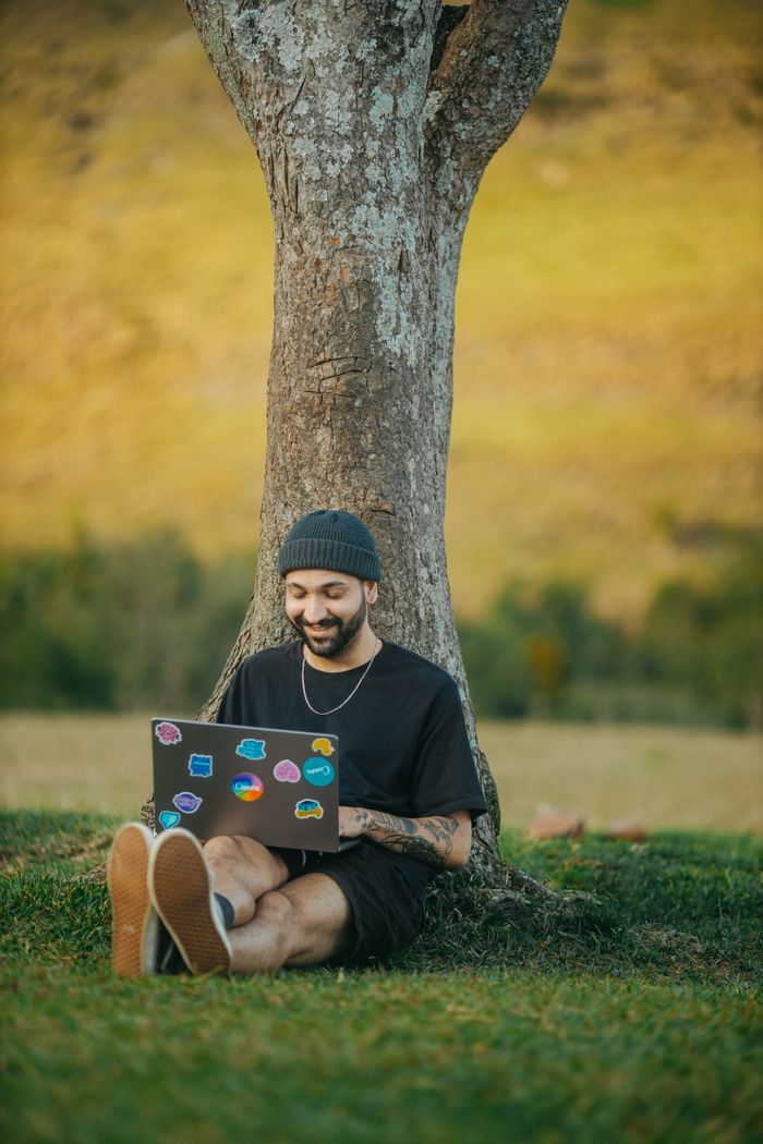 Young man with beard sits under a tree, smiling and using a laptop in a serene meadow.