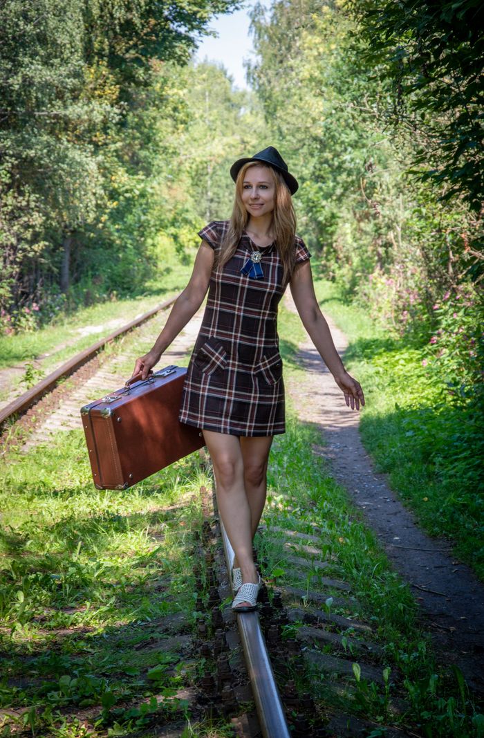 A cheerful woman in a sundress balancing on a railway track with a suitcase in a lush forest setting.
