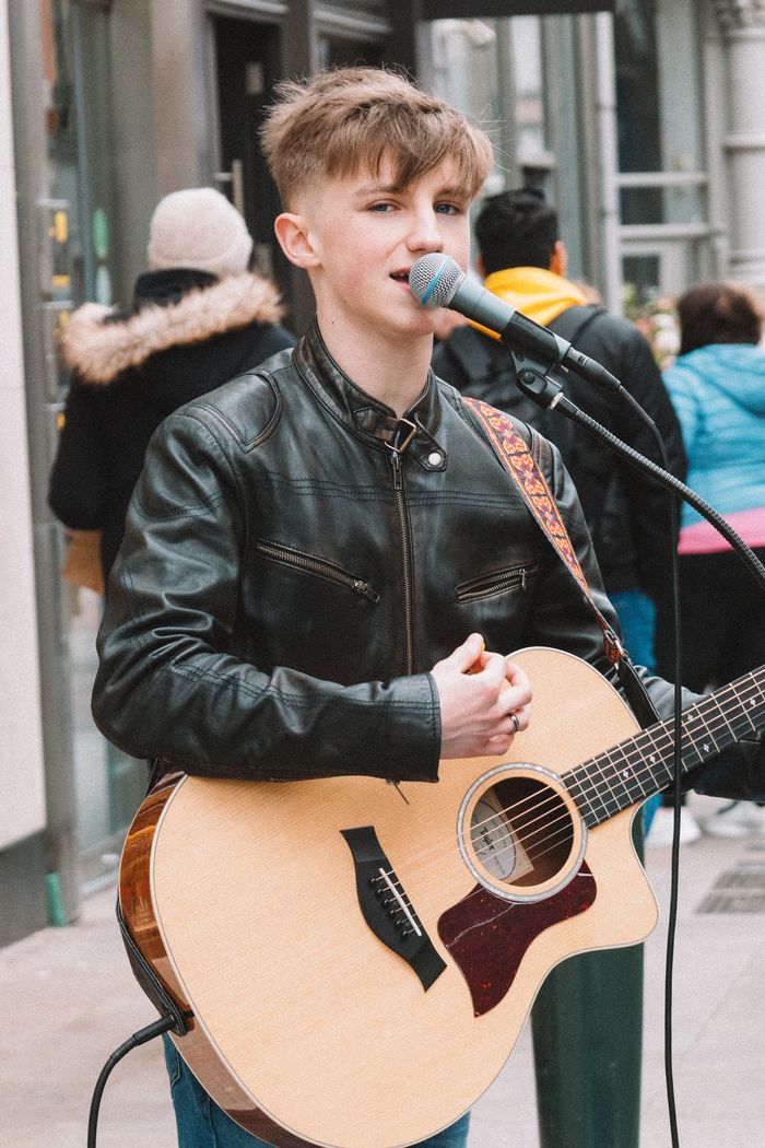 Street musician in a black leather jacket playing acoustic guitar and singing in an urban setting.