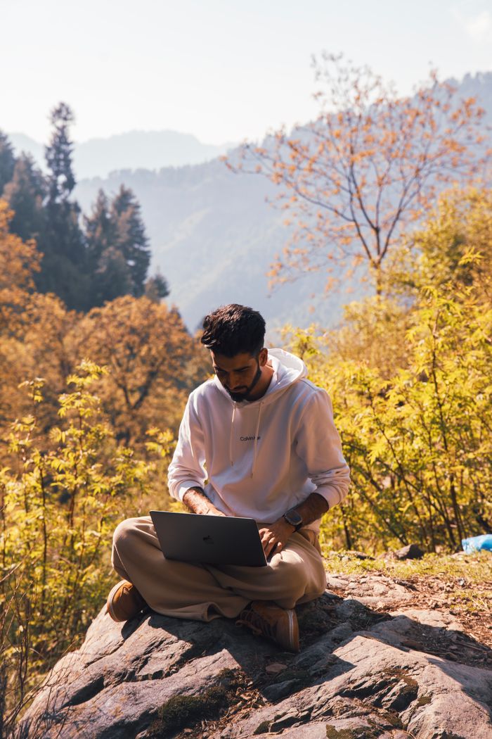 A man sitting on a rock in a forest, using a laptop for remote work in a tranquil natural setting.