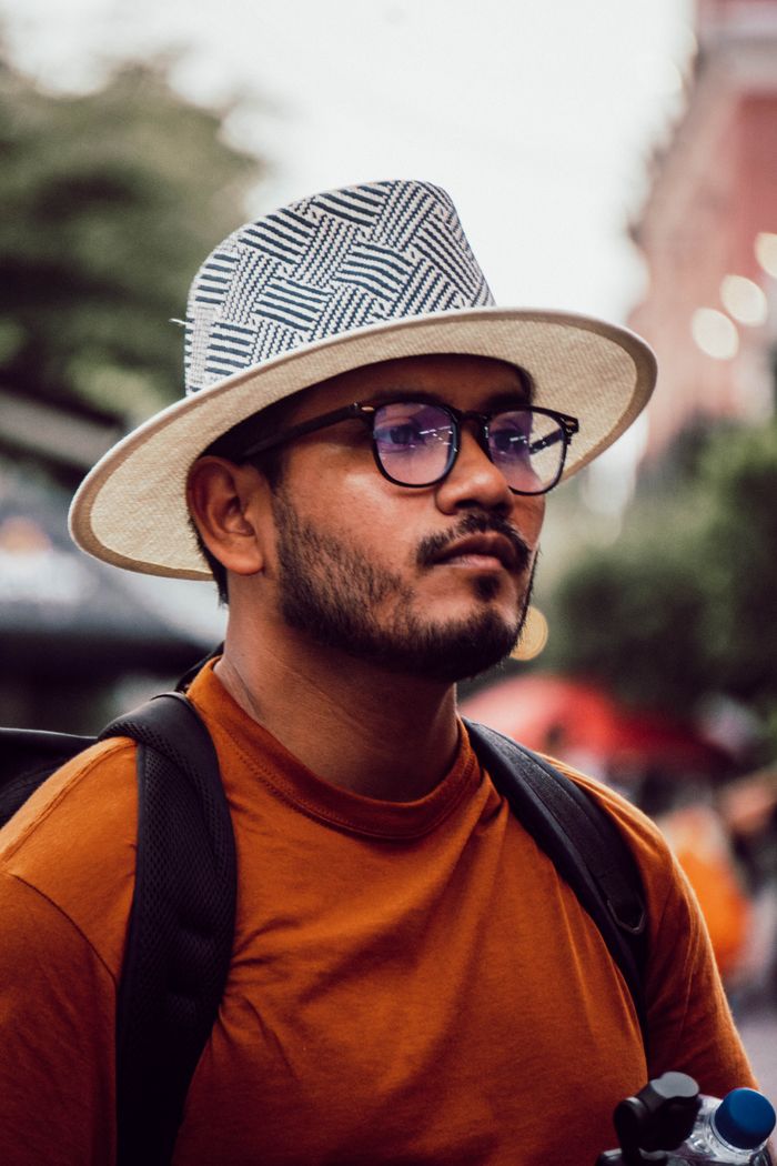 Portrait of a young man outdoors wearing a stylish hat and glasses, with a blurred urban background.